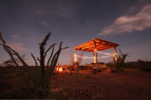 A rustic shelter with string lights under a starry sky, surrounded by desert plants and a campfire.