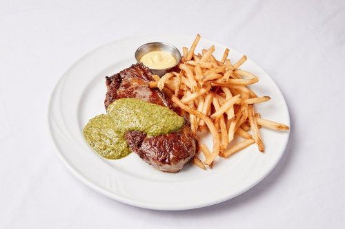 A plate with grilled steak topped with green sauce, served with a side of crispy fries and a small cup of dipping sauce.