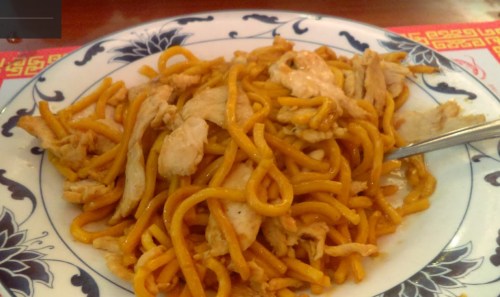 A plate of chicken lo mein with yellow noodles and pieces of chicken, garnished in a decorative blue and white dish.