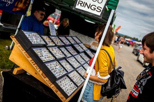 A vendor booth displays numerous rings on a wooden stand, with customers browsing and chatting in an outdoor market setting.