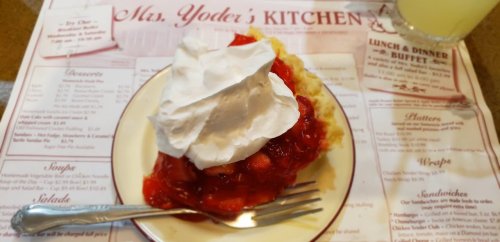 A slice of cherry pie topped with whipped cream on a plate, resting on a menu at Mrs. Yoder's Kitchen.