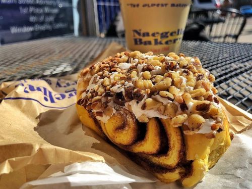 A cinnamon roll topped with nuts and cream cheese frosting, placed on a paper wrapper with a coffee cup in the background.