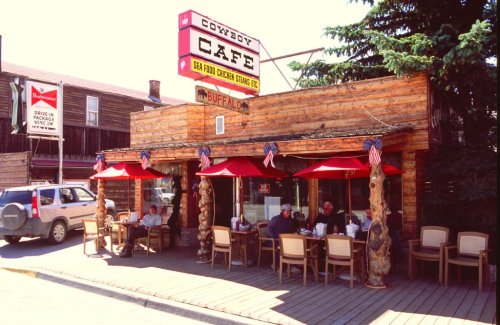 Exterior of Cowboy Cafe with red umbrellas, diners outside, and a rustic wooden facade in a small town setting.