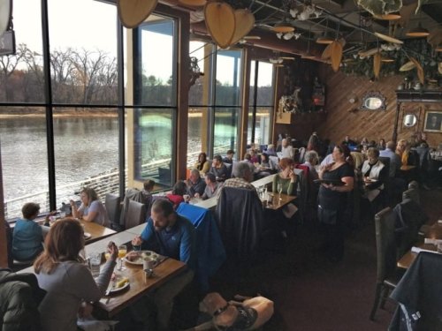 A bustling restaurant with large windows overlooking a river, filled with diners enjoying meals and conversation.