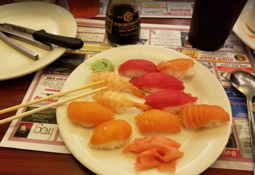 A plate of sushi featuring various types of nigiri, shrimp, and pickled ginger, with soy sauce in the background.