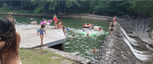 A lively swimming area with children and adults enjoying the water, a wooden dock, and a rocky bank in a green park.
