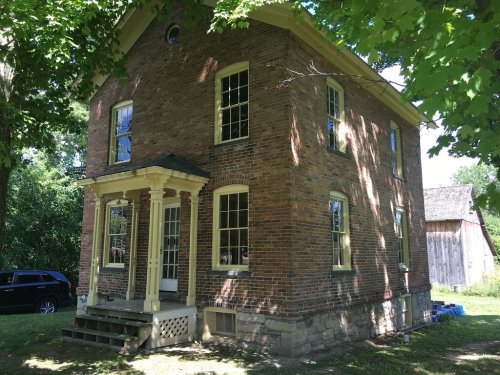A two-story brick house with yellow trim, surrounded by trees and a car parked nearby.