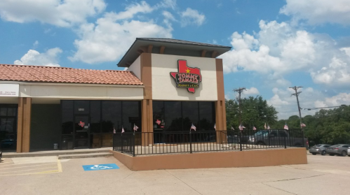 Exterior of a market with a Texas-themed sign, featuring a ramp and flags, under a blue sky with clouds.