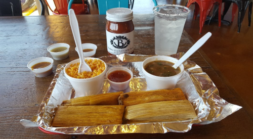 A tray with tamales, rice, beans, sauces, and a drink on a wooden table.