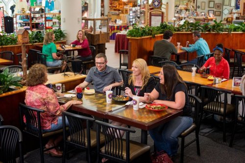 A group of four people enjoying a meal at a bustling food court, surrounded by greenery and other diners.