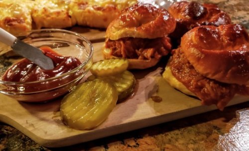 Two pulled pork sandwiches on a wooden board, served with pickles and a small bowl of ketchup.