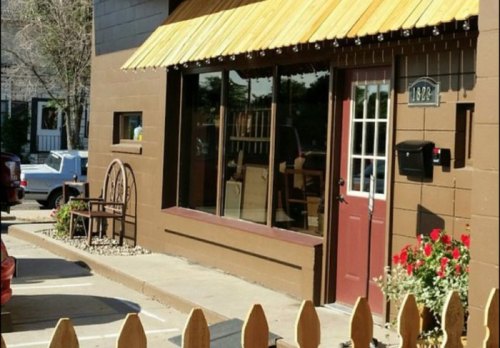 A brown building with a wooden awning, large windows, and a red door, featuring a small garden and seating outside.