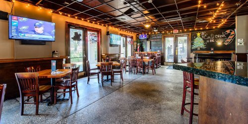 Interior of a restaurant with wooden tables, chairs, a bar, and a TV showing a sports broadcast. Warm lighting and decor.