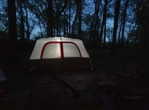 A tent illuminated from within, set in a wooded area at dusk, with trees surrounding it.