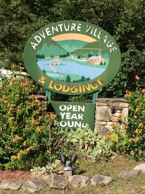 Sign for Adventure Village & Lodging, featuring a scenic landscape and the text "Open Year Round." Surrounded by flowers.