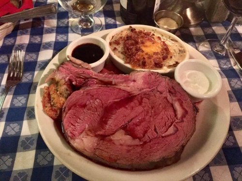 A large serving of prime rib with sides of au jus, creamy potatoes, and a dipping sauce on a checkered tablecloth.