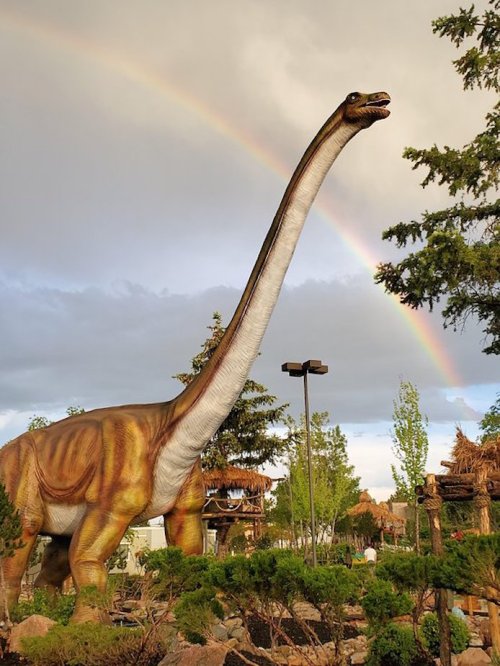 A large dinosaur statue stands under a rainbow, surrounded by trees and greenery.