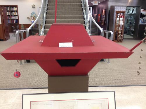 A red, geometric sculpture displayed in a gallery, with a staircase and glass cases in the background.
