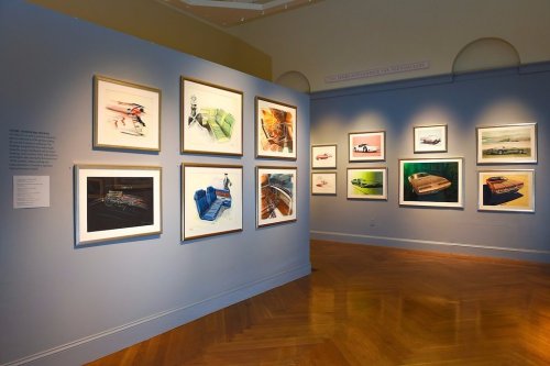 A gallery wall displaying framed automotive sketches and paintings in a well-lit exhibition space.