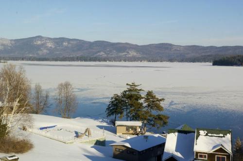 A snowy landscape with a frozen lake, mountains in the background, and houses along the shore under a clear blue sky.