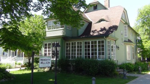 A green Victorian-style house with large windows, surrounded by trees and a sign reading "Meredith Willson."
