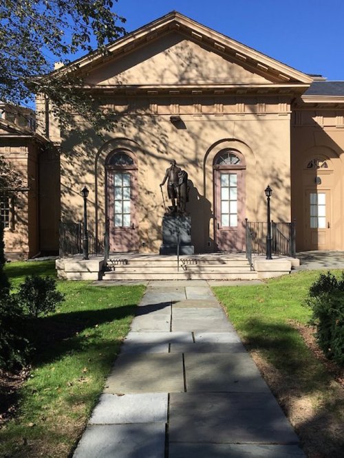 A statue stands in front of a classical building with large windows and a stone pathway surrounded by greenery.