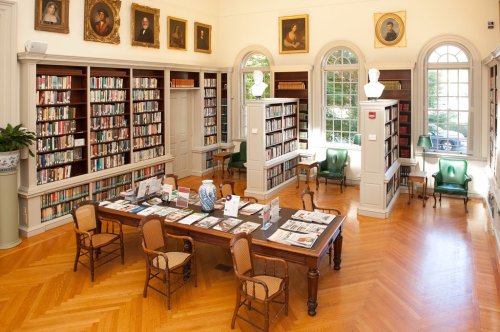 A spacious library with wooden shelves, a large table, and green chairs, featuring portraits on the walls and natural light.
