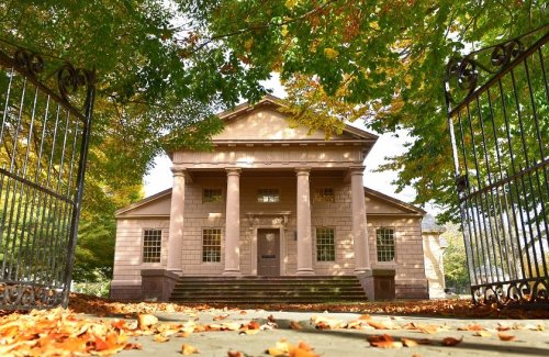 A historic building with columns, framed by trees and autumn leaves, viewed through an ornate gate.