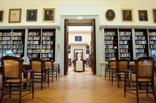 A spacious library with wooden floors, bookshelves, and portraits on the walls, leading to a room with a statue.