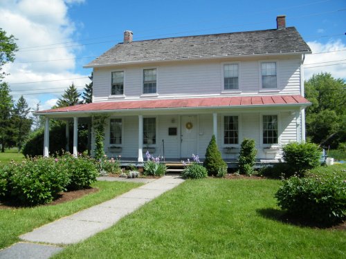 A two-story white house with a red roof, surrounded by green grass and shrubs, under a blue sky with clouds.