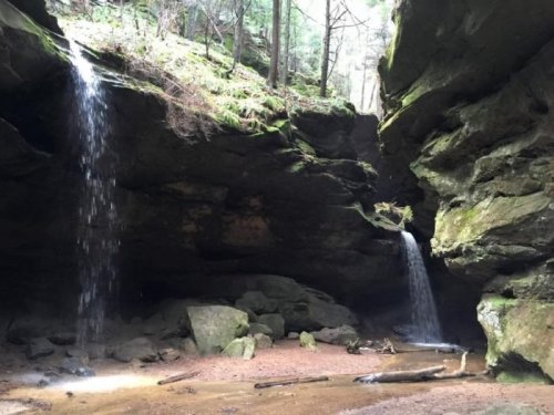 Two waterfalls cascade down rocky cliffs in a lush, green forest, surrounded by moss-covered stones and trees.
