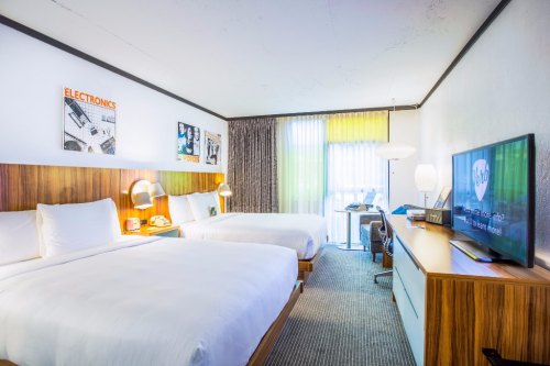 A modern hotel room with two beds, a TV, desk, and large windows with sheer curtains. Decor includes wall art.
