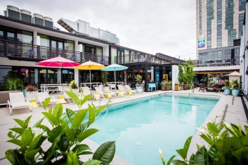 A vibrant pool area with colorful umbrellas, lounge chairs, and greenery, surrounded by modern buildings.
