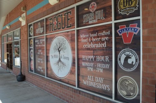 Window display of The White Oak Tavern featuring logos of breweries and a happy hour announcement.