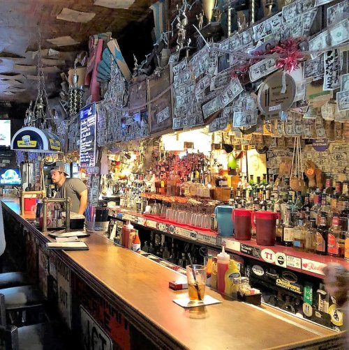 A lively bar scene with a wooden counter, shelves lined with bottles, and dollar bills decorating the walls.
