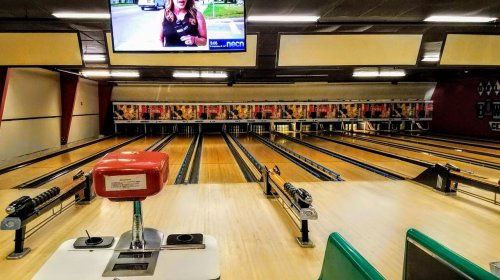 A view of a bowling alley with empty lanes and a TV screen showing a news report in the background.