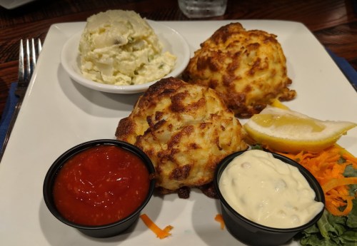 Two golden crab cakes on a plate with lemon, a side of potato salad, and dipping sauces: ketchup and tartar sauce.