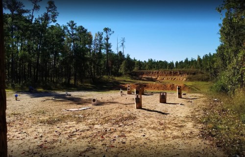A shooting range with targets set up in a wooded area, surrounded by trees and a clear blue sky.