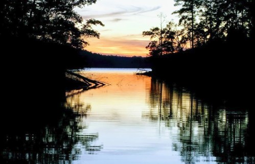 A serene lake at sunset, with trees silhouetted against colorful skies and reflections on the water's surface.