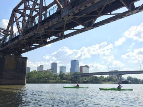 Two kayakers paddle on a river beneath a metal bridge, with a city skyline visible in the background under a blue sky.