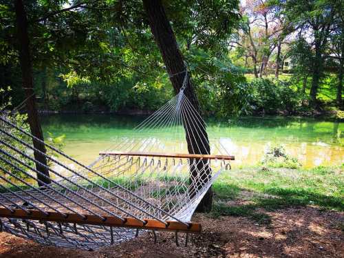 A hammock hangs between two trees by a serene, green river, surrounded by lush foliage.