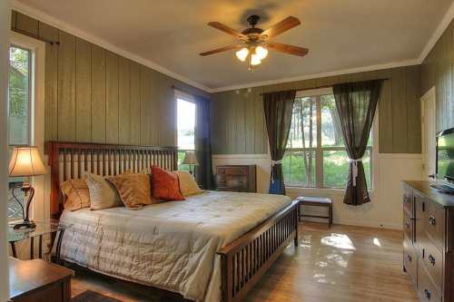 Cozy bedroom with a wooden bed, warm lighting, and large windows overlooking greenery.