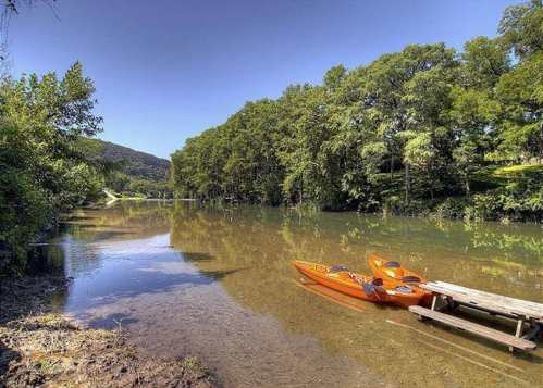 A serene river scene with two orange kayaks docked at a wooden pier, surrounded by lush green trees and clear blue skies.