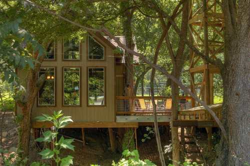 A treehouse with large windows, nestled among trees, featuring a deck and a staircase leading to a higher platform.