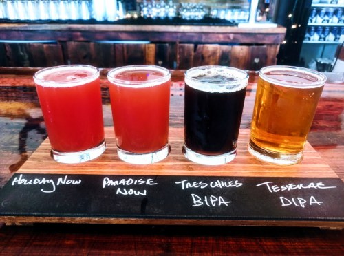 A wooden flight board with four different craft beer samples, labeled with their names, on a bar counter.