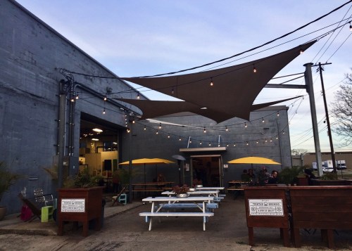 Outdoor seating area with picnic tables, yellow umbrellas, and string lights outside a gray building.