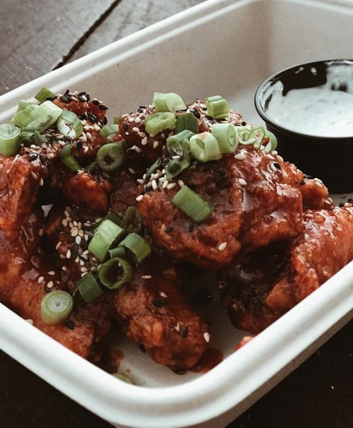 A container of glazed chicken wings topped with green onions and sesame seeds, served with a side of dipping sauce.