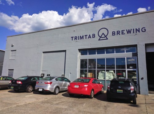 Exterior of TrimTab Brewing with several parked cars and a large window displaying "OPEN" sign. Blue sky with clouds above.