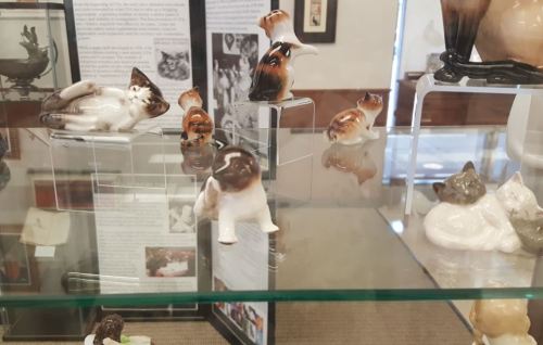 Display of ceramic cat figurines in a glass case, showcasing various poses and colors.