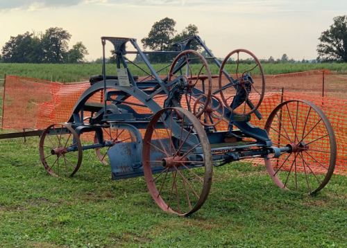 An old blue agricultural machine with large wheels, set against a grassy field and orange safety fencing in the background.